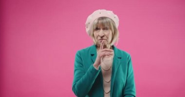 Beyaz tenli emeklilerin portresi. Yaşlı, bereli, ciddi bir kadın. Pembe arka planda tek başına duruyor.