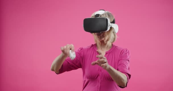 Retrato de cerca de mujer mayor feliz caucásica de pie aislada sobre fondo rosa en gafas VR en auriculares de realidad virtual usando tecnología futurista haciendo gestos con control remoto en el aire — Vídeo de stock