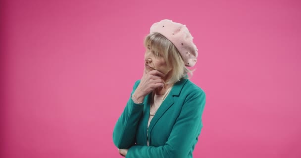 Retrato de pensionista sério caucasiano mulher bonita sênior em boina e casaco verde isolado no fundo rosa com expressão facial pensativa pensando em algum problema para resolver. — Vídeo de Stock