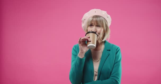 Portré kaukázusi idős szép boldog nő svájcisapkában áll elszigetelt rózsaszín háttér pozitív hangulat gondolkodás álmodik valamit, miközben kezében csésze kávé-to-go és ivás — Stock videók