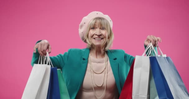 特写快乐快乐的白种人老年女子头戴贝雷帽，站在粉红的玫瑰墙上，在购物后拿着许多购物袋，微笑着，看着相机买方的概念 — 图库视频影像