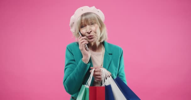 Portret van vrolijke blanke vrolijke stijlvolle oude senior mooie vrouw die staat op rose achtergrond met veel kleurrijke tassen na het winkelen en spreken op oproep op smartphone in een goede positieve stemming — Stockvideo