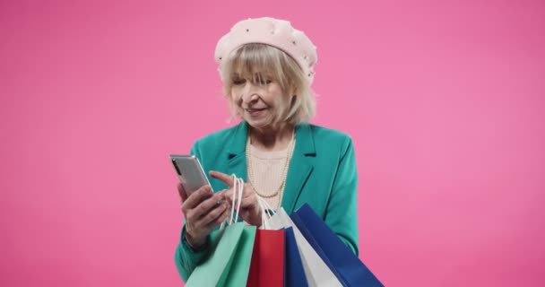 Портрет Кавказької радісної жінки, яка стоїть на рожевому фоні з багатьма барвистими сумками після покупки і смс-перегляду онлайн на смартфоні в позитивному настрої. — стокове відео