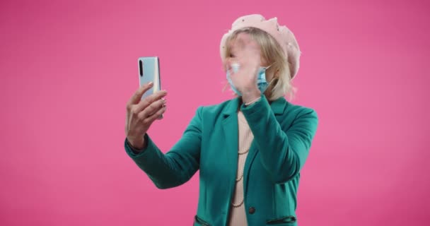 Portrait de belle dame blanche heureuse en béret et masque médical parlant sur appel vidéo sur smartphone isolé sur fond rose. Vieille femme debout sur le mur rose vidéochat sur téléphone portable. — Video