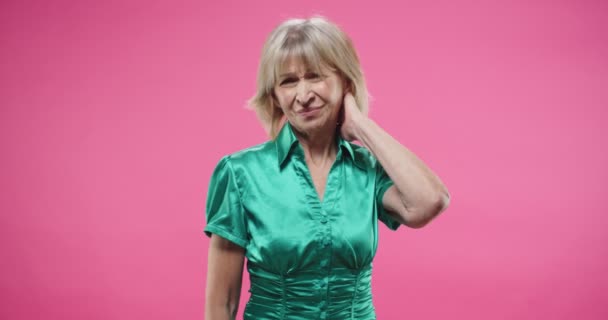 Portrait de triste jolie vieille femme caucasienne en chemisier vert debout sur fond de mur rose sentant la douleur dans le cou faisant massage avec les mains souffrant de sentiments douloureux. Concept de problèmes de santé — Video