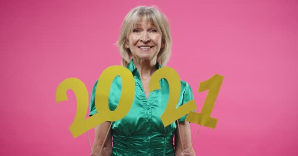 Portrét veselé pozitivní staré bělošky v zelené blůze pózující izolované přes růžové pozadí drží 2021 znamení slaví Nový rok ve sváteční náladě, dívá se na kameru a usmívá — Stock video