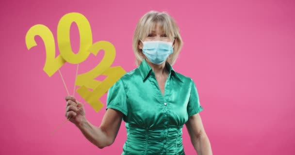 Portrait of Caucasian joyful old Caucasian woman standing over pink background in studio holding 2021 sign looking at camera and smiling, putting medical mask off face and waving hand saying good bye — Stock Video
