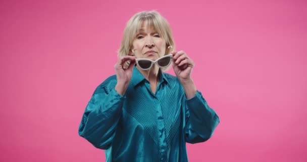 Portrait concept. Caucasian joyful senior pretty stylish female putting on sunglasses while standing on pink background in studio and smiling to camera Elderly happy woman posing isolated on rosy well — Stock Video