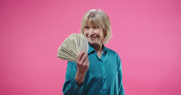 Primer plano retrato de Caucásico senior bastante elegante mujer alegre posando sobre fondo rosa en el estudio, sonriendo de buen humor y mirando a la cámara sosteniendo en la mano muchos dólares. Concepto de dinero — Vídeo de stock