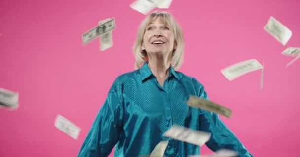 Retrato de mulher caucasiana velho animado alegre em camisa azul posando no fundo rosa de pé em estúdio sob dinheiro chuva dólares caindo de cima olhando para a câmera com emoções felizes positivas — Vídeo de Stock