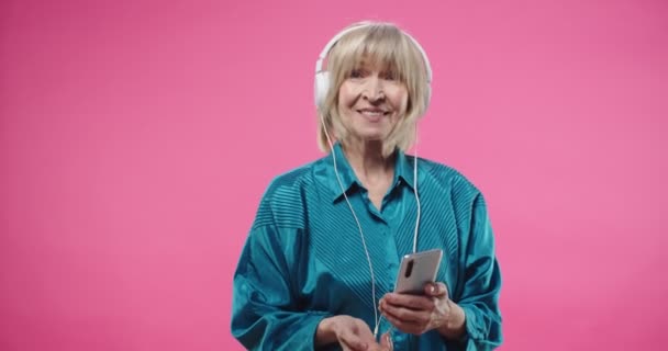 Vit glad söt söt vit senior kvinna lyssnar på musik favorit låt i hörlurar från smartphone med hjälp av och skriva på mobiltelefon, dansa över rosa bakgrund. Fritidsidé — Stockvideo