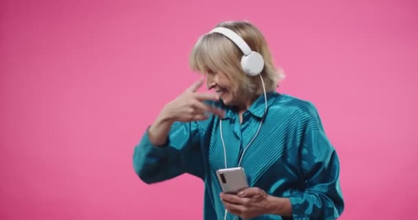 Porträtt av vit glad glad glad glad glad söt gammal kvinna lyssnar på musik favorit låt i hörlurar på smartphone medan du dansar och woving på rosa rosig bakgrund. Kul koncept — Stockvideo