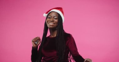 Neşeli, Afro-Amerikalı, Noel Baba şapkalı güzel esmer kadının yakın plan portresi. Stüdyoda pembe arka plan üzerinde komik hareketler yapıyor. Mutlu bir ruh hali içinde.