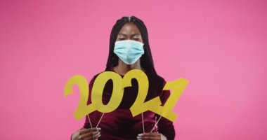 Güzel, Afro-Amerikan neşeli, tıbbi maskeli genç bir kadın 2021 'de karantinada kutlama yaparken pembe arka planda duruyor. Yeni yıl kutlaması. Portre konsepti