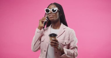 Portrait of happy African American young beautiful woman in sunglasses speaking on smartphone, having good talk receiving some shocking funny news and holding cup of coffee standing on pink background