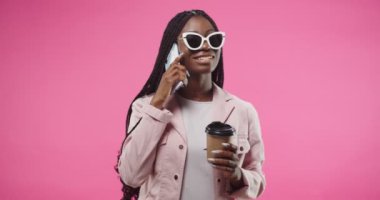 Portrait of cheerful pretty African American young woman wearing sunglasses chatting on cellphone, having nice talk with funny face expression and holding cup of coffee in hand standing on pink wall