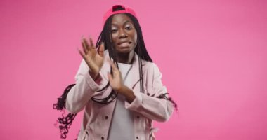 Portrait of African American young beautiful cheerful woman wearing red cap in positive mood funny moving and dancing making gestures with hands celebrating win, expressing joy, isolated on rosy wall