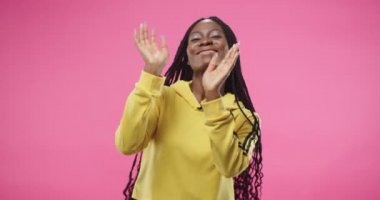 Sarı kapüşonlu tişörtlü, neşeli, güzel, Afro-Amerikan bir kadının portresi. Mutlu bir şekilde dans ediyor, komik hareketler yapıyor ve elleri pembe arka planda duruyor.