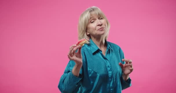 Portrait of cheerful pretty Caucasian senior blonde lady in blue shirt holding in hand donut eating and enjoying tasty delicious food standing on pink wall in studio with funny face expression. — Stock Video