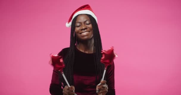 Nahaufnahme Porträt einer fröhlichen jungen hübschen Afroamerikanerin mit Weihnachtsmütze, die tanzt und Gesten wie Trommeln macht, die kleine Luftballons auf rosa Hintergrund halten, um Neujahr zu feiern — Stockvideo