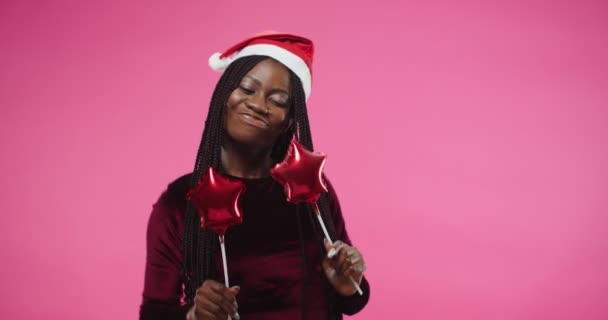 アフリカ系アメリカ人は、手のスティックに小さな風船でサンタの帽子のダンスで若い美しい女性を喜び、新年を祝うピンクの壁にお祝いの気分で楽しんで移動します。肖像画を閉じる — ストック動画