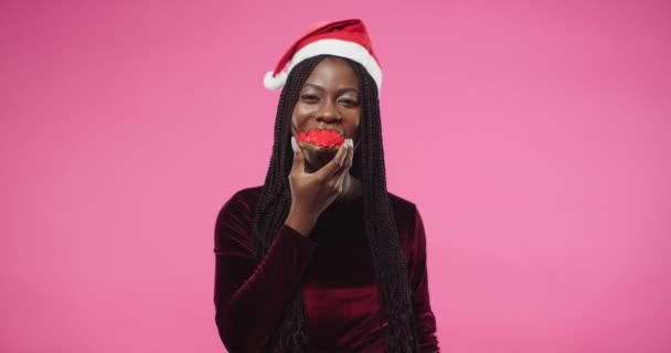 Primer plano retrato de la joven afroamericana alegre hermosa mujer de pie posando sobre la pared rosa en el estudio comiendo sabrosa galleta en Año Nuevo. Celebración 2021. Cara feliz. Emociones positivas — Vídeo de stock