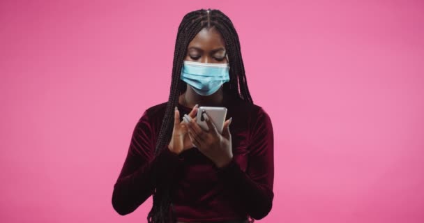Retrato de afroamericana hermosa joven mujer seria con máscara médica se para en el estudio sobre fondo rosa rosado y mensajes de texto, escribiendo navegar por Internet en línea en el dispositivo del teléfono inteligente — Vídeos de Stock