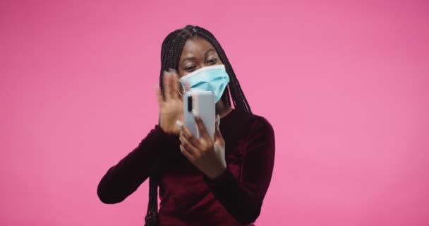 Portret van vrolijke Afro-Amerikaanse mooie jonge vrouw in medisch masker staan op roze achtergrond roze muur en videochatting spreken op video oproep online op smartphone-apparaat — Stockvideo