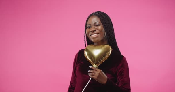 Close up portrait of happy smiling young African American female in positive mood holding in hand golden heart shape balloon and sending air kiss while standing on rosy pink wall and looking at camera — Stock Video