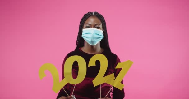 Gros plan portrait de jolie afro-américaine joyeuse jeune femme portant un masque médical et tenant dans les mains 2021 signe célébrant le nouvel an en quarantaine tout en se tenant debout sur fond rose. — Video