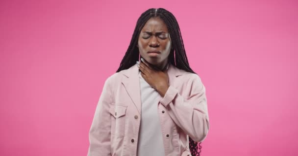 Portret afro-american frumos tânăr rău trist de sex feminin stă pe fundal de perete roz în studio având senzație dureroasă în gât având probleme de sănătate. Femeia bolnavă suferă de durere — Videoclip de stoc