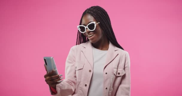 Porträt einer fröhlich lächelnden jungen hübschen Afroamerikanerin in rosafarbener Jacke und Sonnenbrille, die isoliert auf rosigem Hintergrund steht und auf einem Online-Videotelefon mit dem Smartphone telefoniert und mit der Hand wedelt — Stockvideo