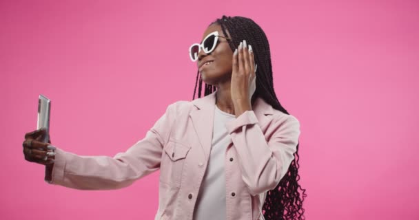 Portrait of happy cheerful young beautiful African American woman in pink jacket and sunglasses posing isolated on rosy background taking selfie photos on smartphone, photoshoot on cellphone — Stok video