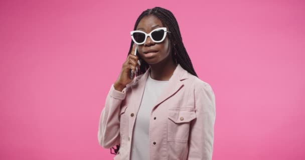 Портрет радостной счастливой африканской молодой красивой девушки в солнечных очках, болтающей по телефону, получающей шокирующие новости с удивленным выражением лица, стоя на розовой стене — стоковое видео