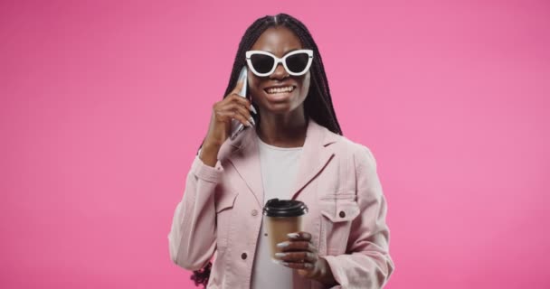 Portrait of African American young beautiful joyful female in sunglasses calling on smartphone, speaking having good talk with friend and drinking coffee while standing over pink background in studio — Stok video