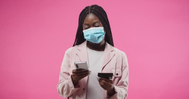 Portrait of African American young joyful pretty woman in mask typing on smartphone paying with credit card making purchase on internet standing on pink background. E-commerce. Shopping online — Stock Video