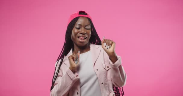 Close up retrato de alegre bela jovem afro-americana positiva em vermelho boné dançando alegremente fazendo movimentos engraçados e gestos com as mãos enquanto posando sobre fundo rosa. Conceito divertido — Vídeo de Stock
