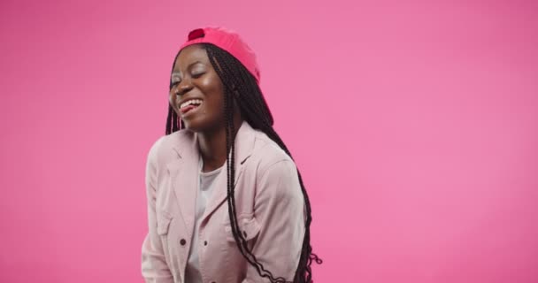 Retrato de la alegre joven afroamericana bonita mujer con gorra roja se encuentra aislado sobre el fondo rosa en el estado de ánimo positivo posando a la cámara haciendo movimientos divertidos y expresión de la cara. Concepto modelo — Vídeos de Stock
