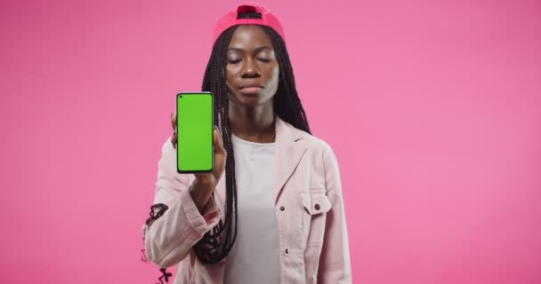 Retrato de una joven afroamericana bastante seria con gorra roja está aislada sobre fondo rosa mirando a la cámara y sosteniendo en la mano el teléfono inteligente con pantalla verde. Celular con llave de croma — Vídeos de Stock