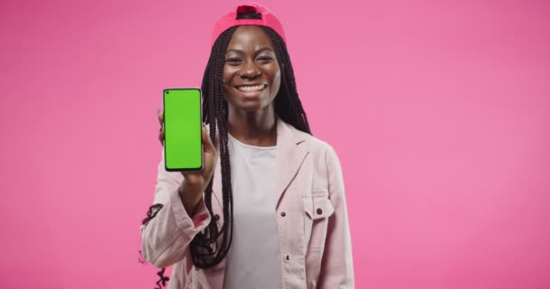 Afroamericana bastante feliz joven con gorra roja se encuentra aislada sobre fondo rosa con sonrisa en la cara mirando a la cámara y sosteniendo el teléfono inteligente con pantalla verde Celular con croma clave Retrato — Vídeos de Stock