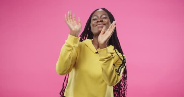 Retrato de alegre feliz bela jovem afro-americana positiva em camisola com capuz amarelo dançando alegremente fazendo movimentos engraçados e gestos com as mãos em pé sobre fundo rosa — Vídeo de Stock