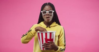 Pembe arka planda izole edilmiş patlamış mısır kutusunu tutan Afro-Amerikalı genç ve mutlu bir kadının portresi. Sarı kapüşonlu kadın patlamış mısır yiyor ve gözlüklü film izliyor. Kadın film izliyor.