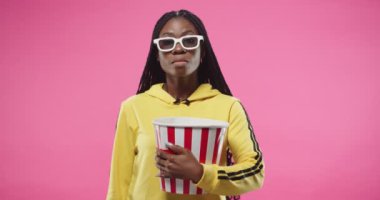 Portrait of concentrated serious African American young beautiful woman standing isolated on pink wall background, holding popcorn box in hands and watching movie or film in glasses. Leisure concept