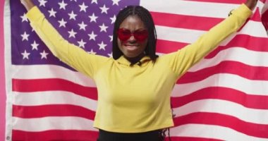 Kırmızı gözlüklü, sarı kapüşonlu, Amerikan bayrağını taşıyan ve gülümseyen güzel Afro-Amerikan genç esmer kadın pembe duvar arkasında tek başına duruyor. Portre