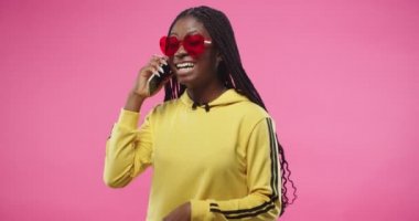 Afro-Amerikan genç, neşeli, kırmızı gözlük takan, kalp şeklinde pembe arka planda izole edilmiş, cep telefonuyla konuşan, gülen ve gülümseyen güzel bir kadın. Portre konsepti