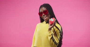 Sarı kapüşonlu ve kırmızı gözlüklü Afro-Amerikalı güzel bir kadın pembe arka planda yalnız poz veriyor müziğe doğru dans ediyor ve elinde bisküvi kekiyle komik bir gülümseme oluşturuyor. Portre