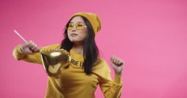 Sarı gözlüklü, müzik ya da şarkıyla dans eden genç Asyalı güzel bir kadının portresi stüdyodaki pembe duvarda duran altın kalp şeklindeki balonu ritmik bir şekilde tutarak eğleniyor.