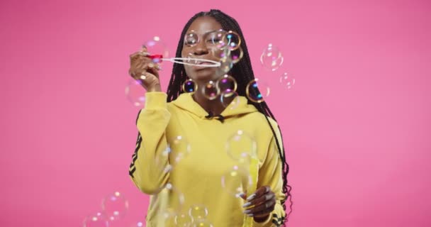 Pretty Afro-Amerikaanse jonge vrolijke glimlachende vrouw in gele sweatshirt blazen zeepbellen in positieve stemming terwijl staan op roze achtergrond Portret van mooie vrouw in studio Fun concept — Stockvideo