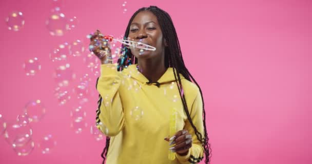 Retrato de la hermosa mujer joven afroamericana alegre con capucha amarilla que sopla burbujas de buen humor mientras está de pie en la pared de fondo rosa y sonriendo. burbuja de jabón. Concepto de ocio — Vídeos de Stock