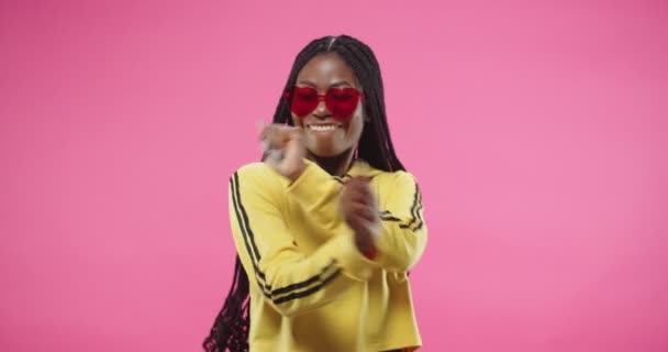 Retrato de una joven afroamericana positiva bastante feliz hembra con capucha amarilla en forma de corazón bailes de gafas modernas haciendo movimientos rítmicos a la música aislada sobre fondo rosa. Concepto de bailarina — Vídeos de Stock